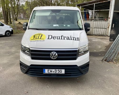 Weißer Volkswagen Transporter mit einem „Deufrains“-Logo auf der Vorderseite, geparkt im Freien in der Nähe eines Gebäudes und von Bäumen.